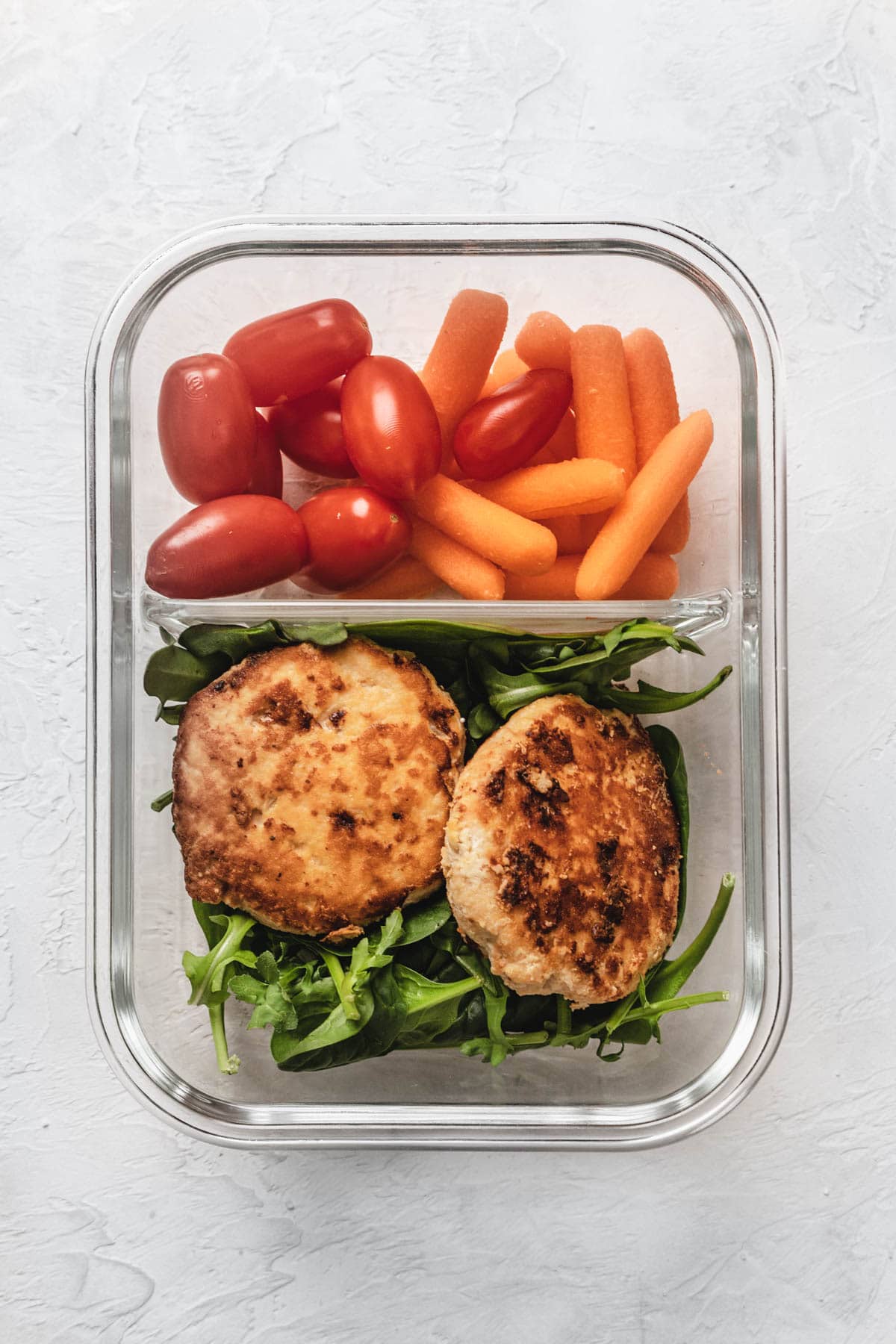 a meal prep container with tomatoes, carrots, chicken burgers, and lettuce