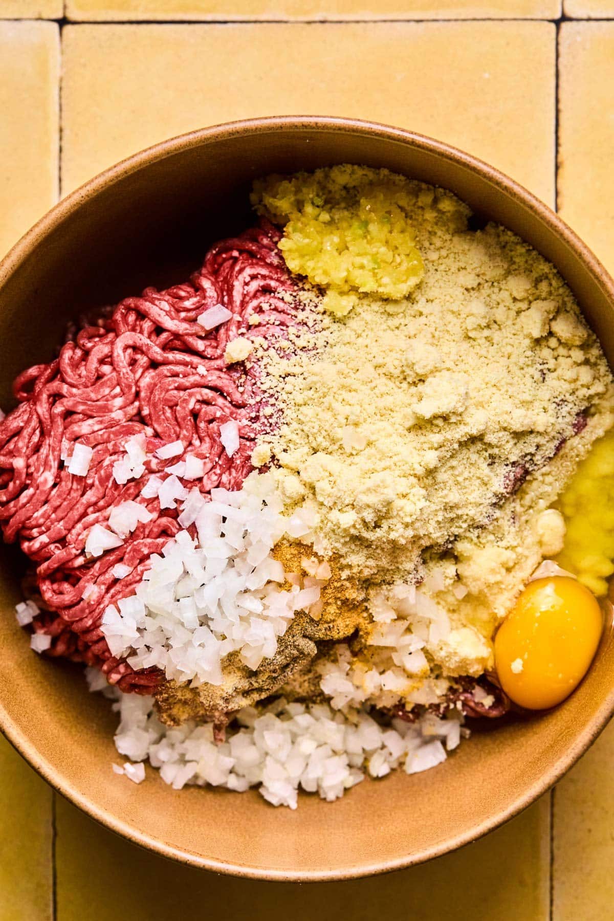 A large bowl with ground beef, egg, almond flour, choppd onion, coconut aminos, garlic, ginger, salt, and pepper.