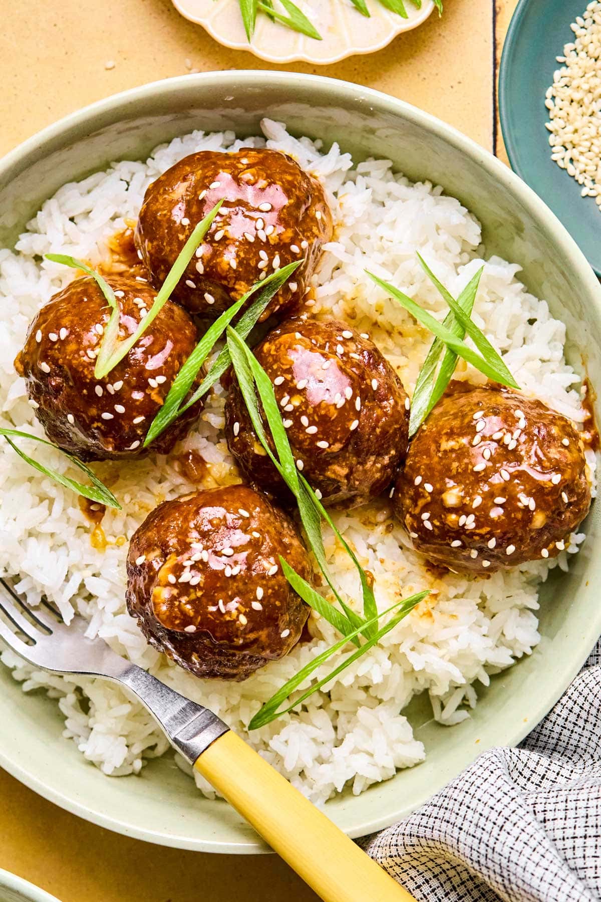 Asian meatballs coated in a sticky sauce garnished with green onions and sesame seeds over a bed of rice.