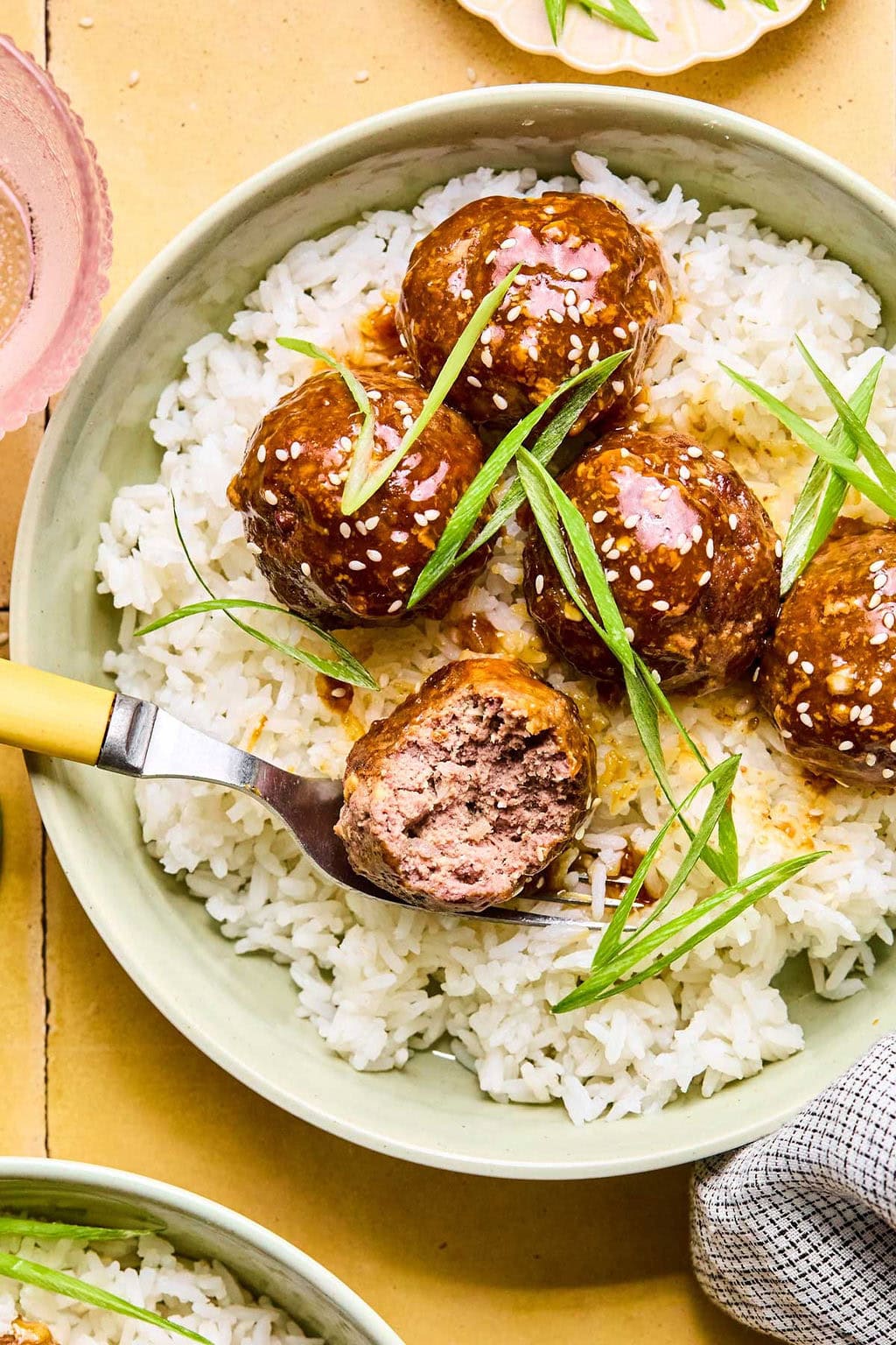A close-up of a green bowl of Asian meatballs garnished with green onions and sesame seeds over a bed of rice. One meatball has a bite taken out of it.