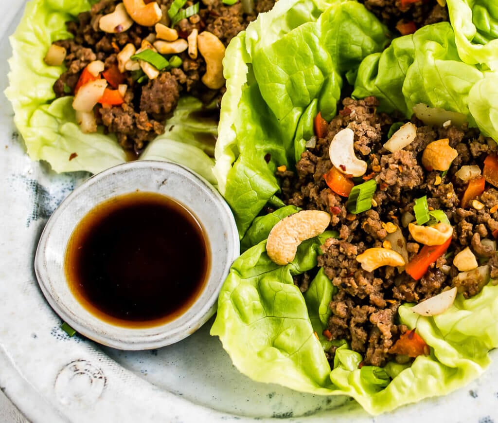 closeup of lettuce wraps filled with ground beef, red peppers, cashews, and green onions next to a small bowl of sauce. 