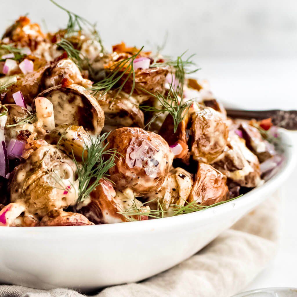 Potato salad in a white bowl with bacon, ranch, red onion and fresh dill. 