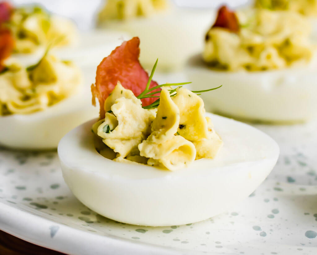A closeup picture of a deviled egg garnished with fresh dill and topped with crispy bacon.