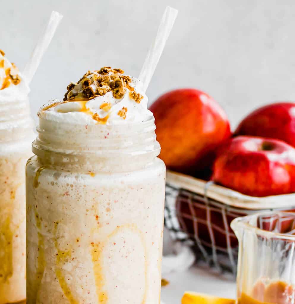 Up close shot of a Caramel Apple Smoothie with whipped cream, caramel sauce and granola with apples in the background