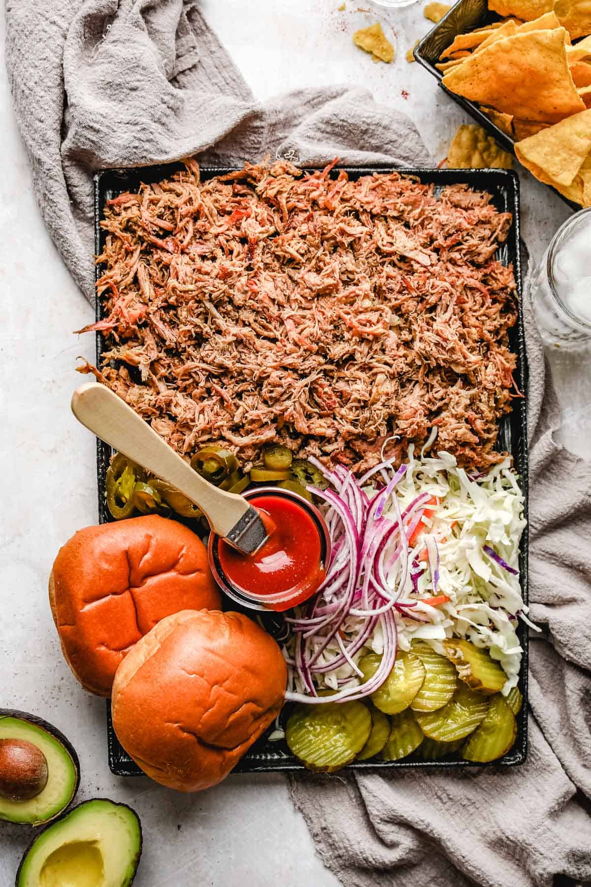 a sheet pan filled with shredded smoked pork shoulder, jalapenos, hamburger buns, sliced onions, pickles, coleslaw and bbq sauce 