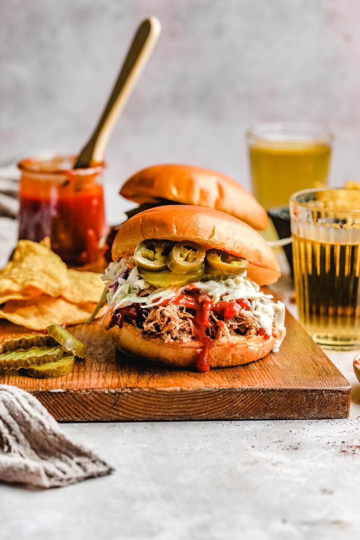 two traeger pulled pork sandwiches on a wooden cutting board next to a pile of tortilla chips, a jar of bbq sauce, and a few sliced pickles