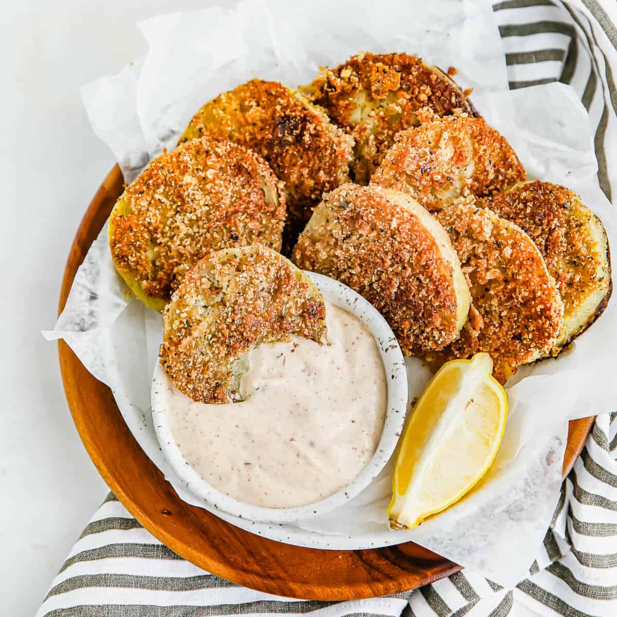 Keto fried green tomatoes on a round wooden serving platter with a small nesting bowl of Mac's awesome sauce. One of the tomatoes is dipped in the sauce.