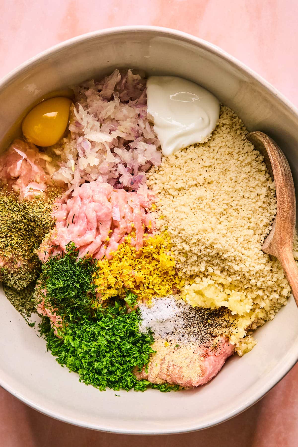 A bowl filled with ingredients including a raw egg, yogurt, minced onions, breadcrumbs, ground meat, chopped herbs, lemon zest, salt, pepper, and spices. A wooden spoon rests on the side.