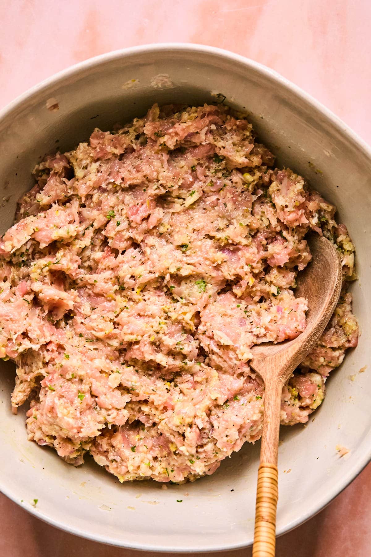 A bowl filled with seasoned raw ground meat mixture, containing visible herbs and small bits of ingredients. A wooden spoon rests in the bowl, indicating mixing or preparation. The surface beneath is a soft pink color.