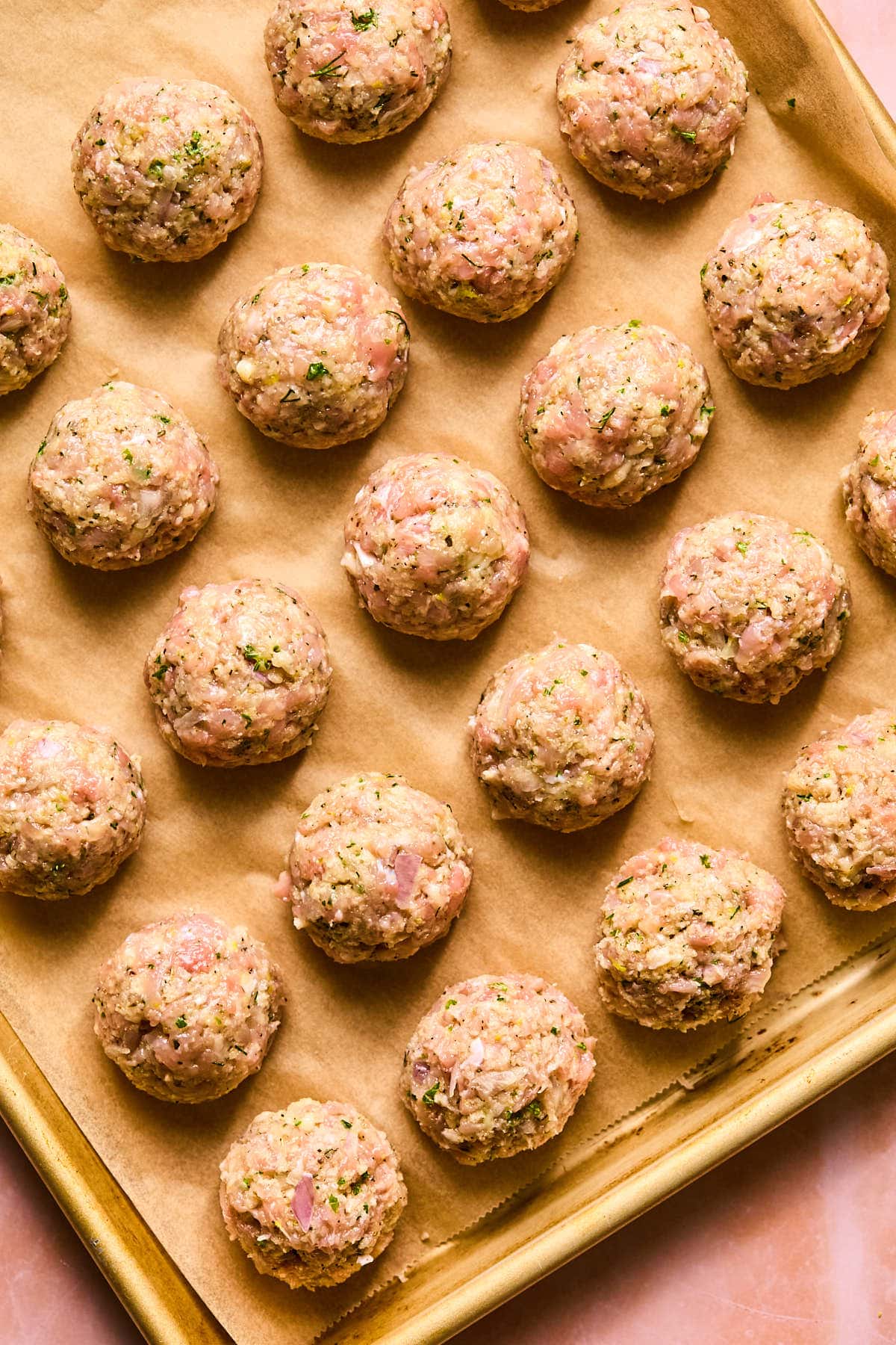Raw meatballs arranged in neat rows on a parchment-lined baking sheet. The meat mixture contains visible herbs and seasonings, giving texture to the surface of each round meatball. The backdrop is a warm, pinkish hue.