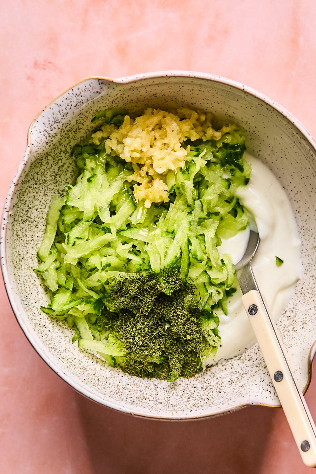 A bowl with grated cucumber, minced garlic, dried dill, and yogurt about to be mixed, placed on a pink surface. A spoon rests inside the bowl.