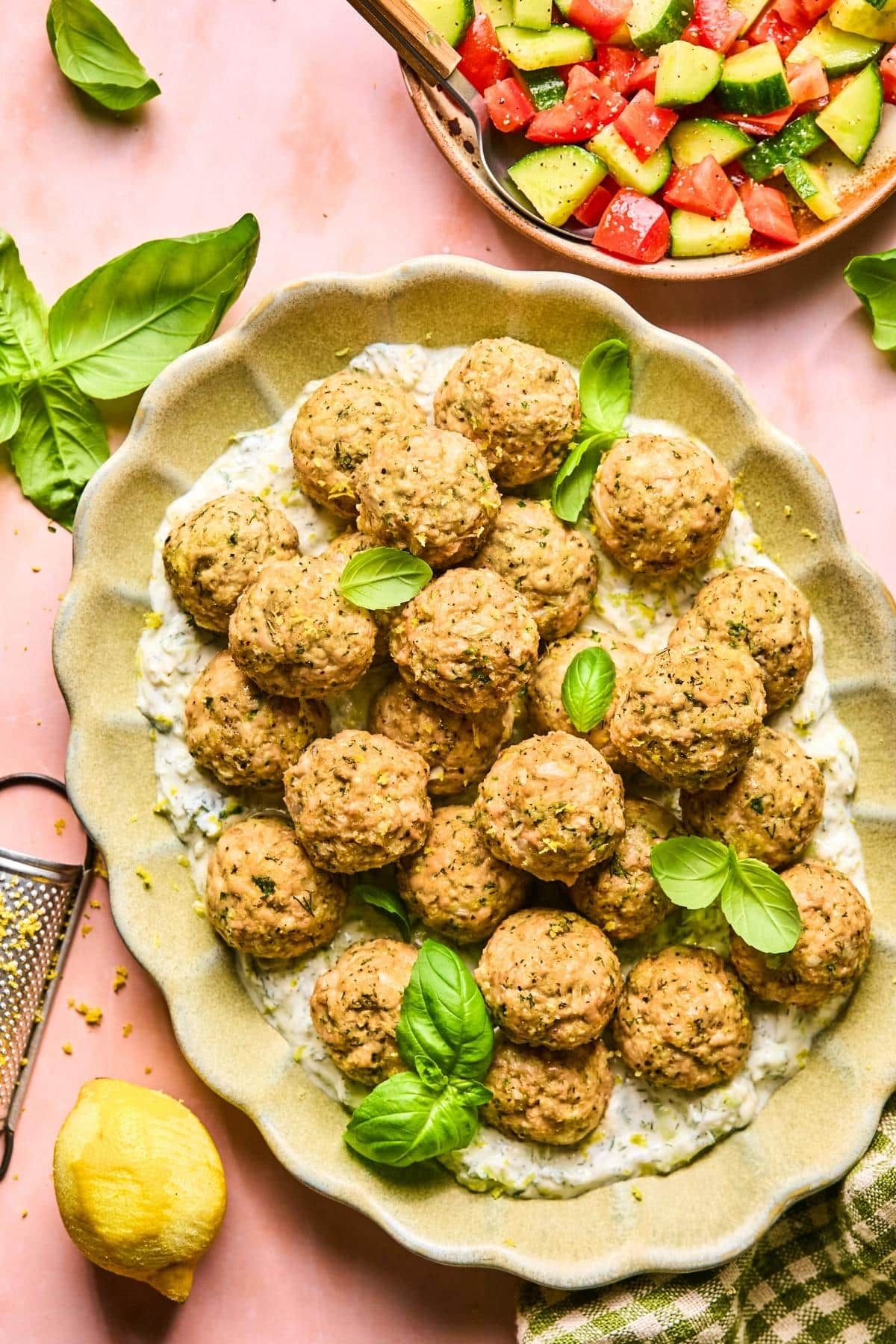 A platter of Greek chicken meatballs garnished with fresh basil leaves. The meatballs rest on a bed of creamy white sauce. A side salad with diced cucumbers and tomatoes is visible in the top corner. A lemon and grater are nearby.