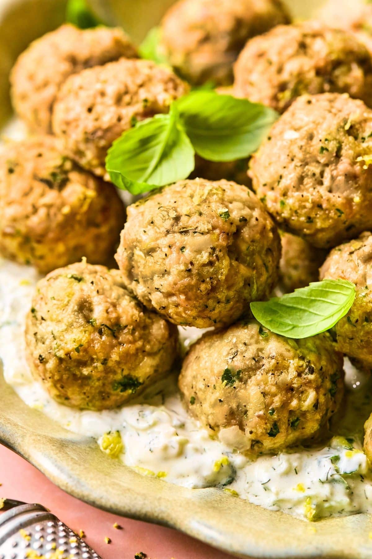 A plate of baked Greek chicken meatballs garnished with fresh basil leaves, served over a creamy sauce with herbs. The meatballs have a golden-brown crust and appear seasoned with herbs and spices.
