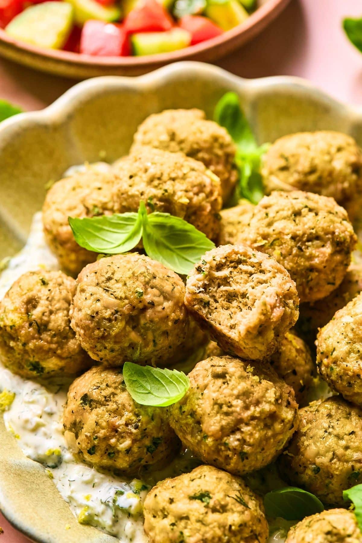 Golden-brown meatballs stacked on a plate with a creamy white sauce and garnished with fresh basil leaves. A bowl of colorful chopped vegetables is partly visible in the background.