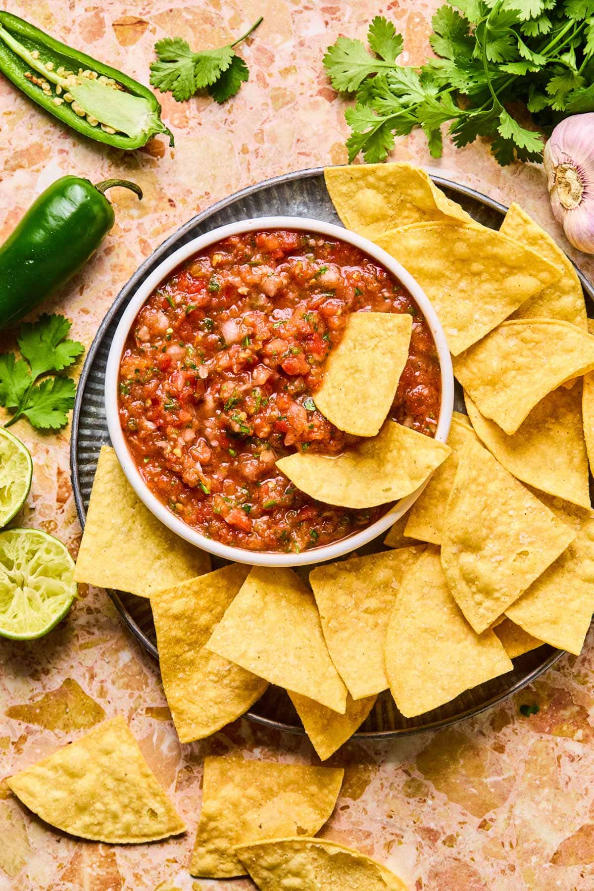 Bowl of vibrant garden fresh salsa with diced Roma tomatoes, onions, jalapeños, and cilantro, served with tortilla chips on a warm toned stone-like surface.