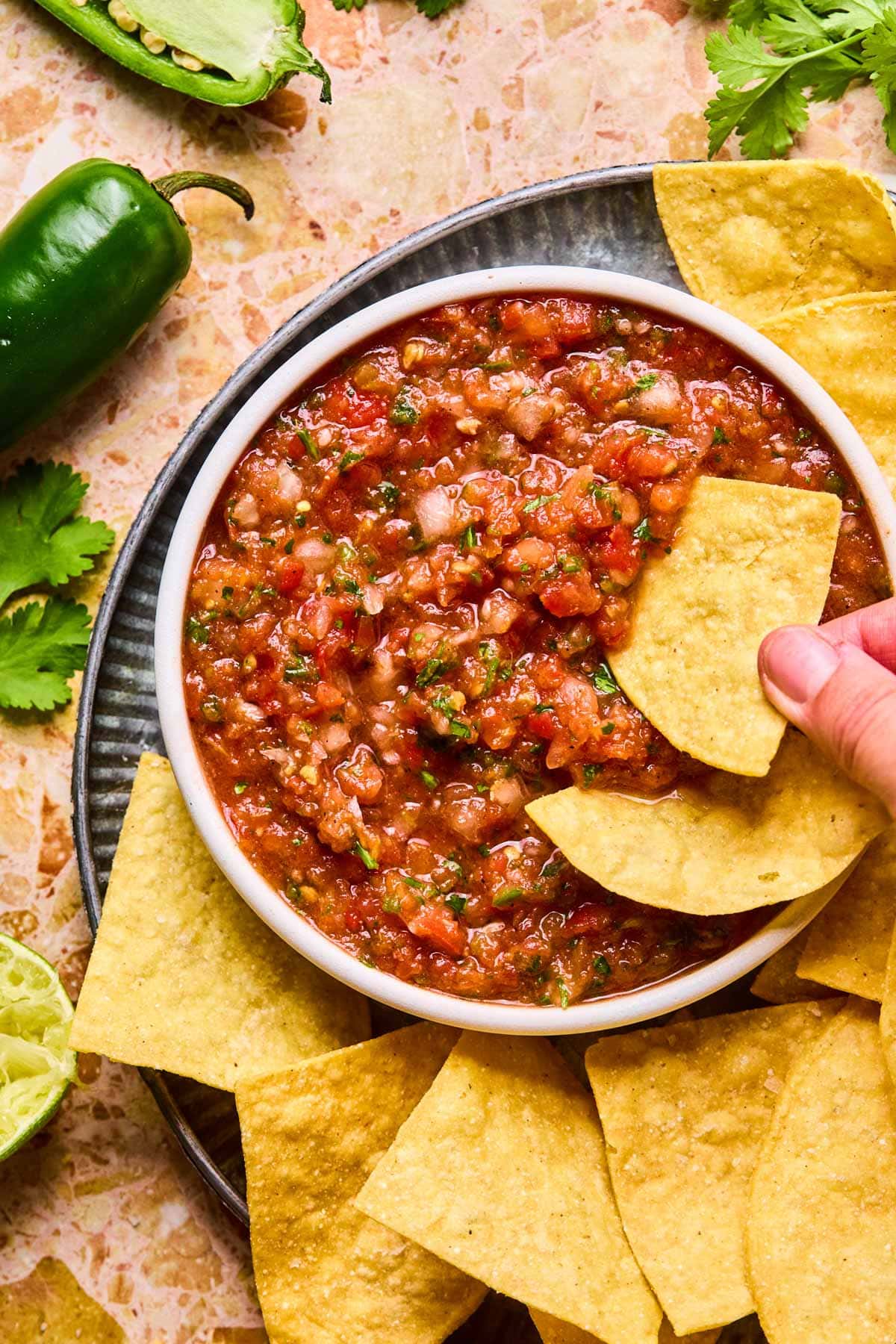 Close-up of chunky blended salsa in a white bowl, with a hand dipping a tortilla chip in it.