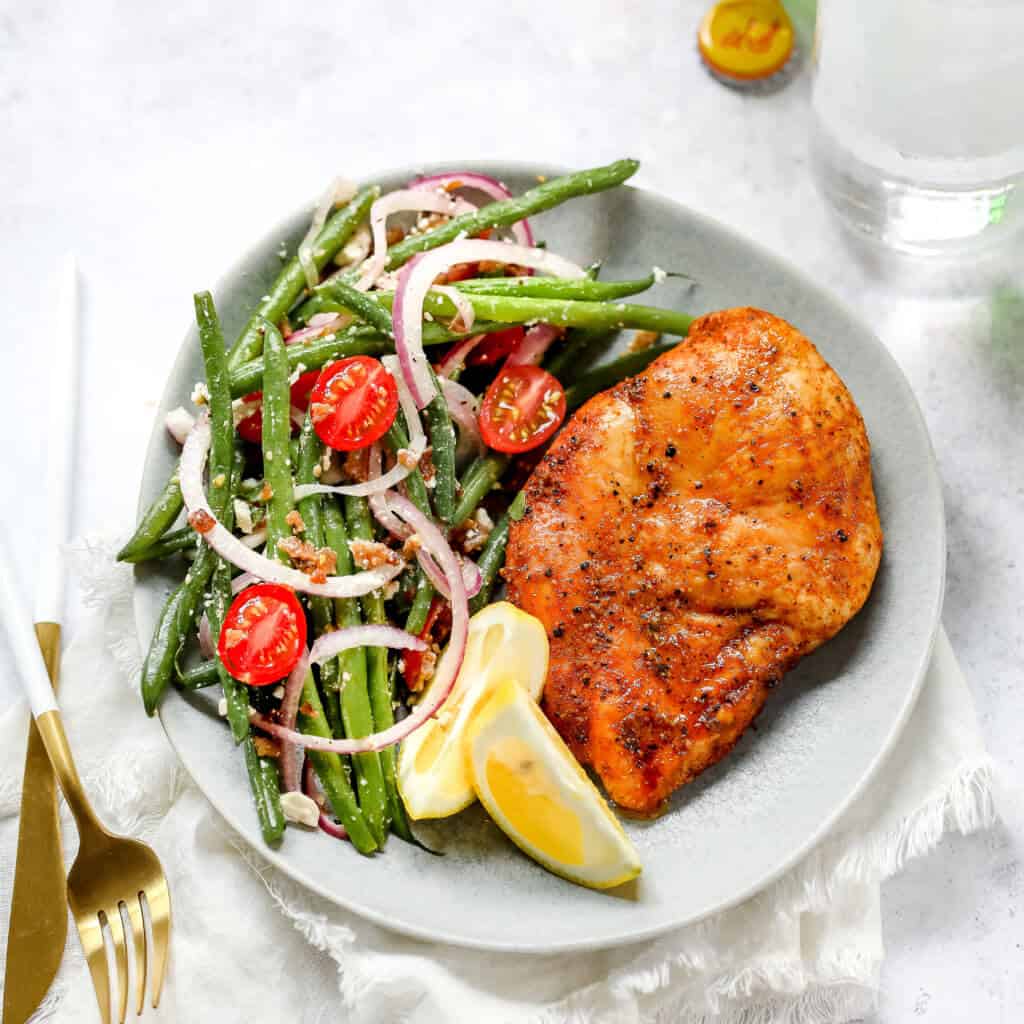 A photo of a plate with a chicken breast, lemon slices and green bean salad