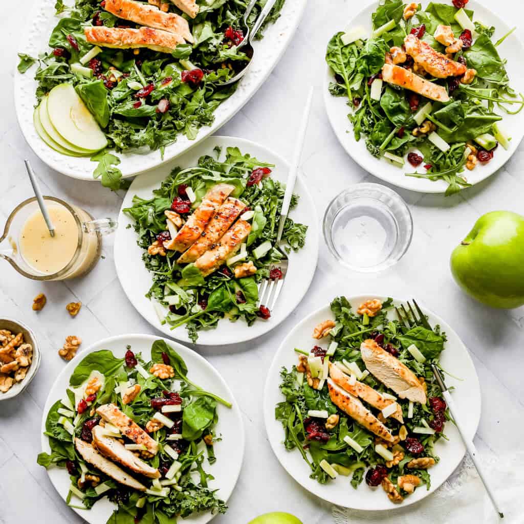 Harvest Chicken Apple Salads on five white plates, a glass jar filled with Honey Lemon Vinaigrette and a spoon, an empty glass cup, a green apple, and a bowl of walnuts with a white background