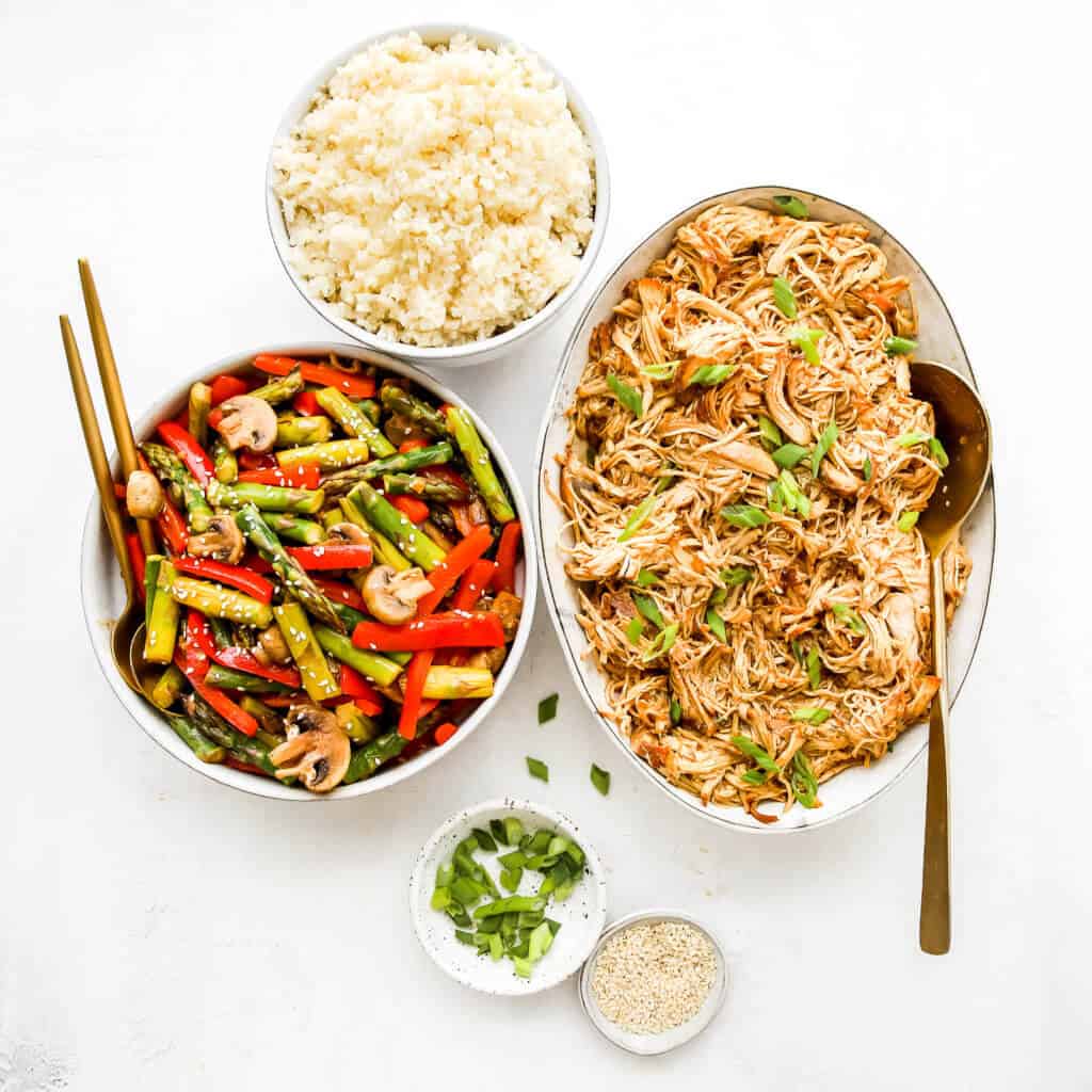 cauliflower rice, veggies, and shredded chicken in white bowls with gold spoons and a white background. 