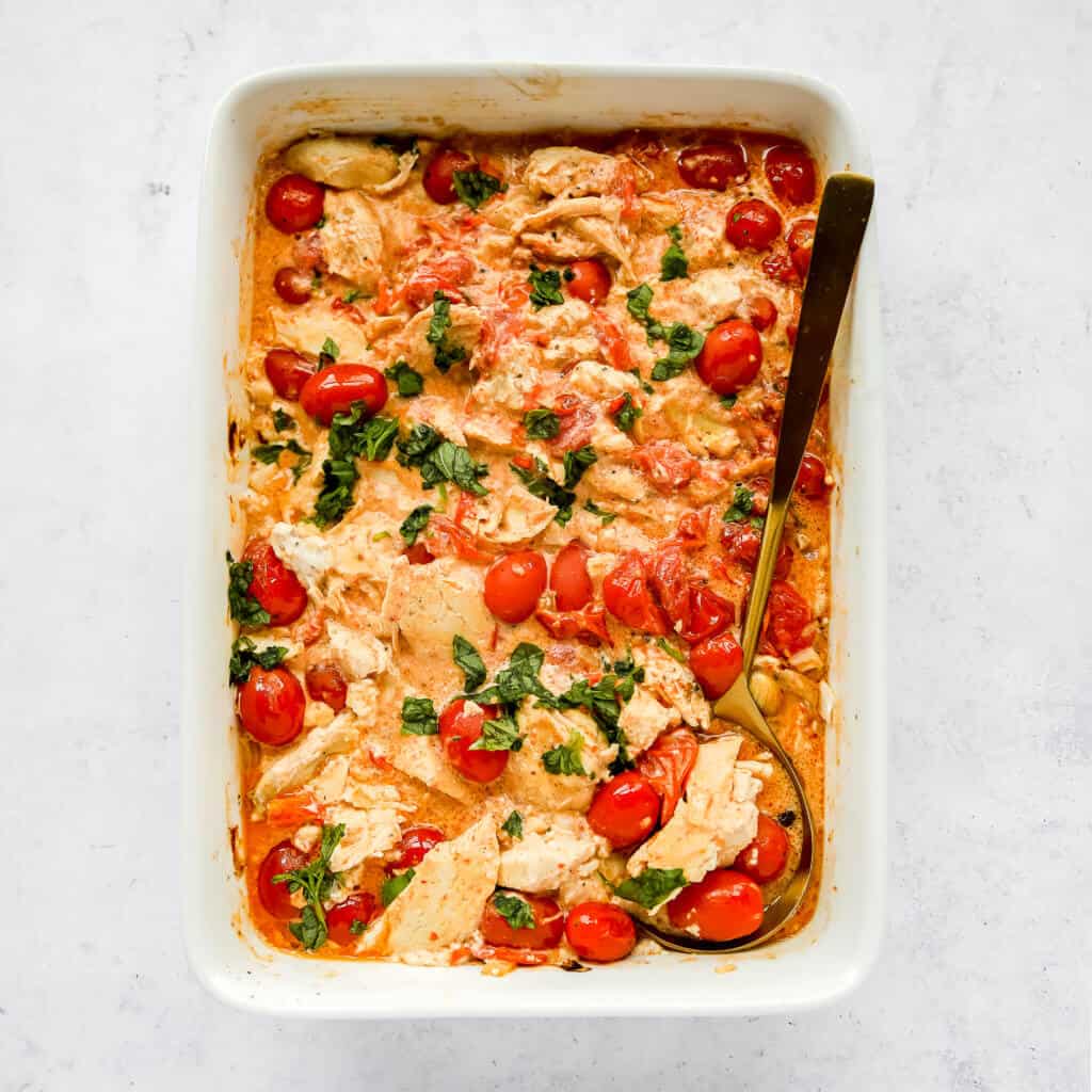 creamy chicken and tomato bake topped with basil in a white dish with a gold spoon and a white background