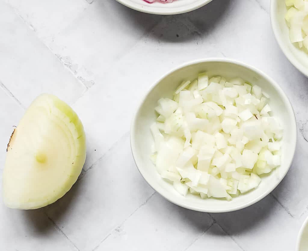 half of a yellow onion next to diced yellow onion in a white bowl with a white background