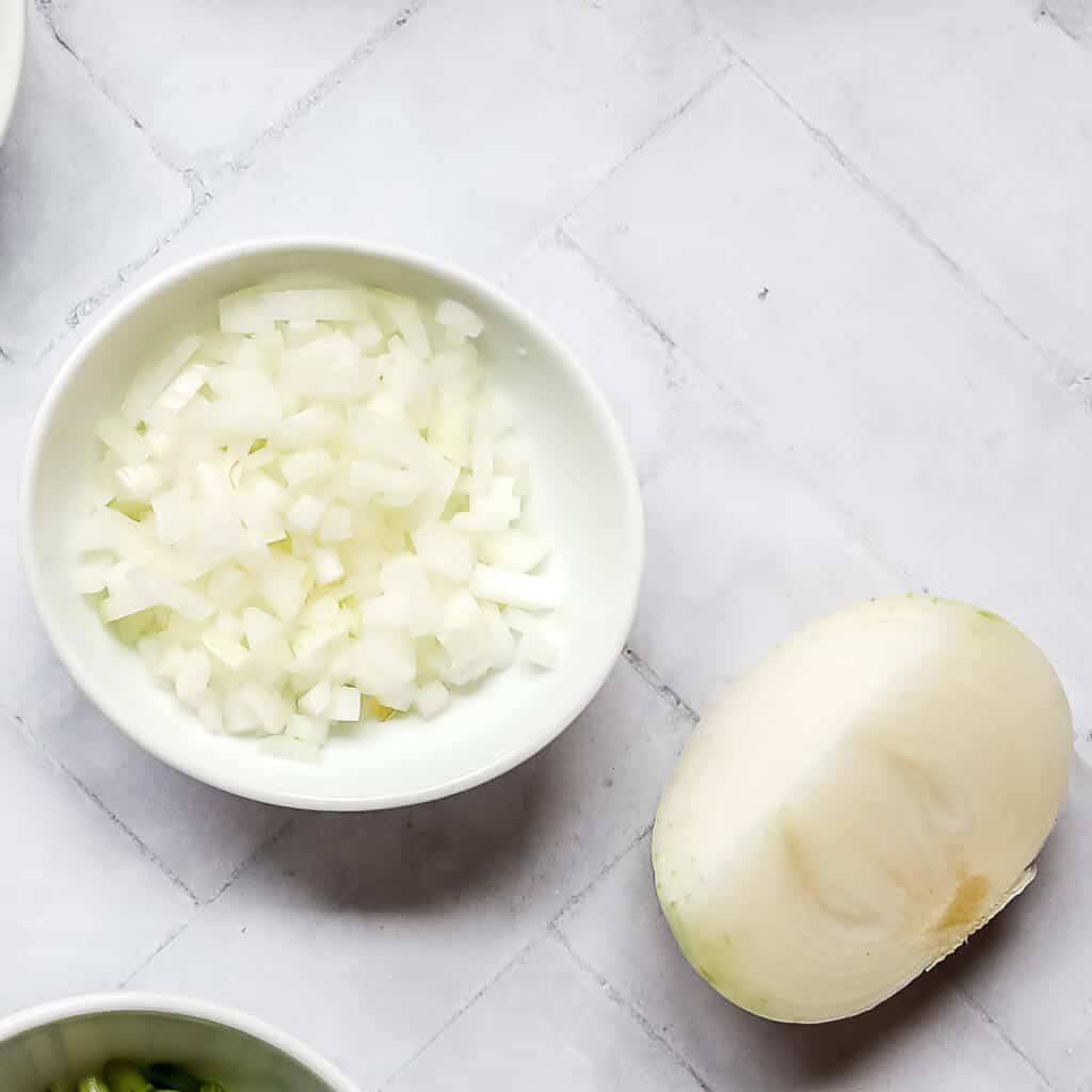 half of a white onion next to diced white onion in a white bowl with a white background