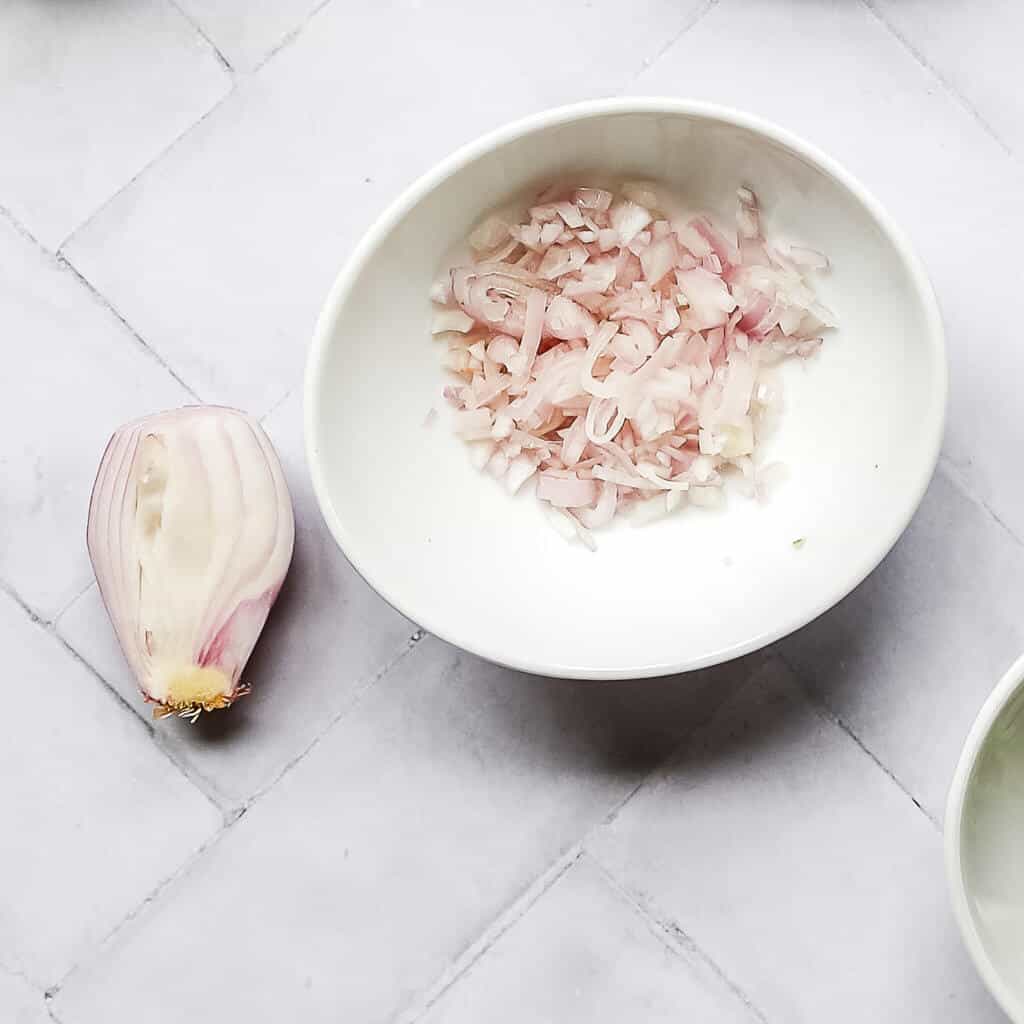 half of a shallot next to diced shallots in a white bowl with a white background