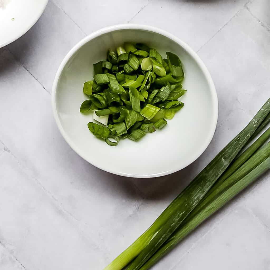 bunch of green onion next to diced green onion in a white bowl with a white background