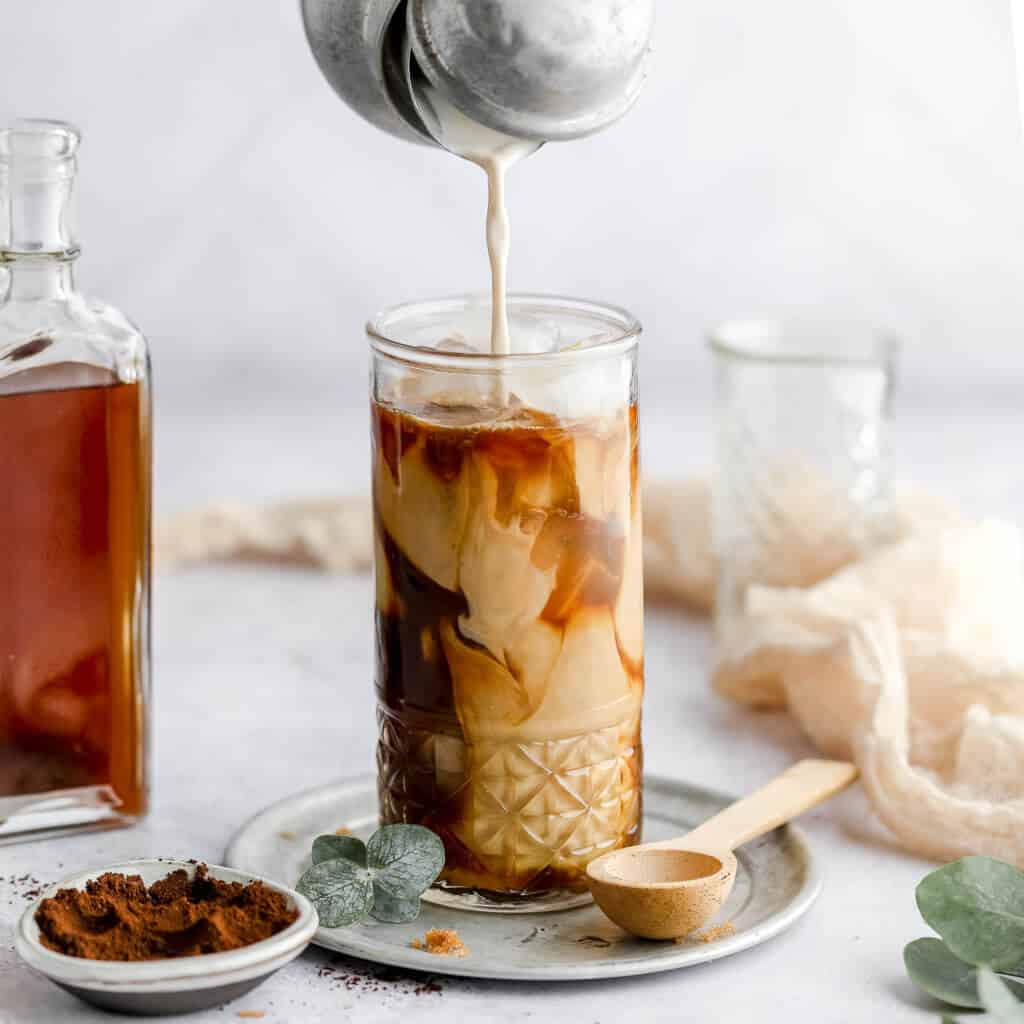 keto brown sugar latte in a tall glass with a silver carafe pouring almond milk into the glass, a glass bottle filled with brown sugar syrup next to the glass, and a pinch bowl filled with cinnamon with a white background.