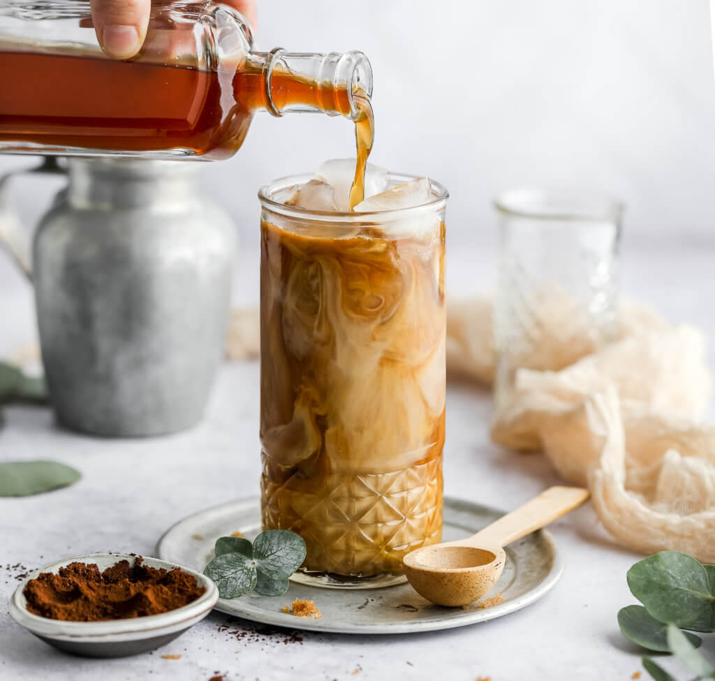 keto brown sugar latte in a tall glass with a glass bottle pouring brown sugar syrup into the glass, a silver carafe next to the glass, and a pinch bowl filled with cinnamon with a white background.