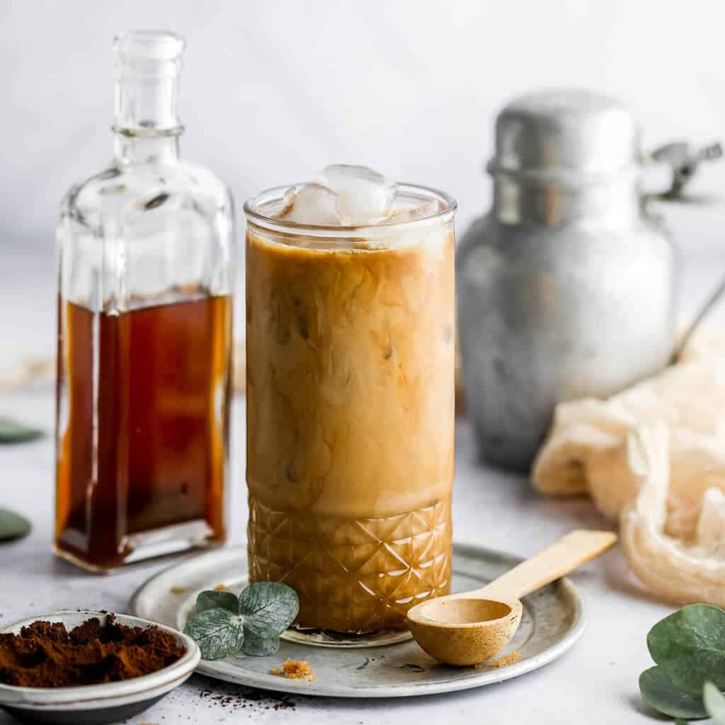 keto brown sugar latte in a tall glass next to a glass bottle filled with brown sugar syrup, a silver carafe next to the glass, and a pinch bowl filled with cinnamon with a white background.