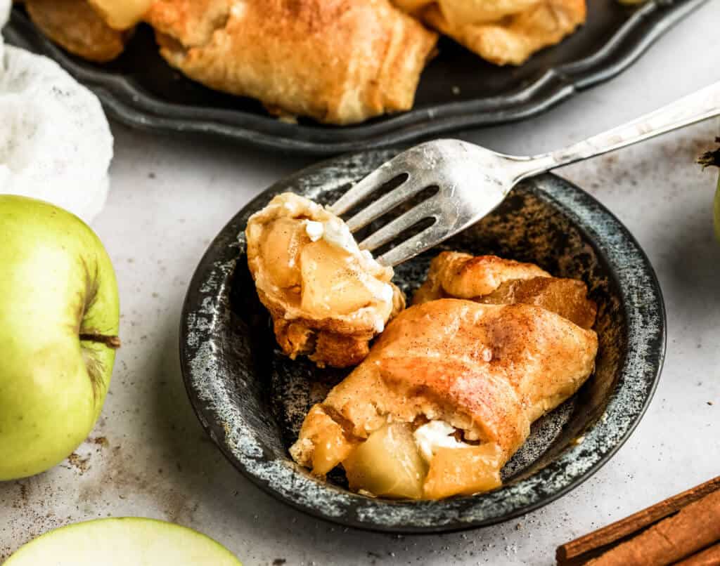 a silver fork picking up a bite of a crescent roll apple pie that is stuffed with apple pie filling and cream cheese and sprinkled with cinnamon sugar in a black bowl.