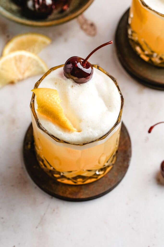 egg white topped whiskey sour cocktail garnished with a cherry and orange rind in a high ball glass.
