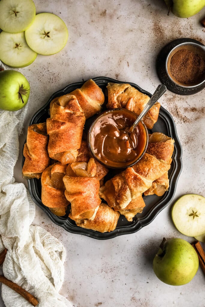 a bunch of mini apple pies made with crescent rolls on a black serving tray with a bowl of caramel dipping sauce in the middle.