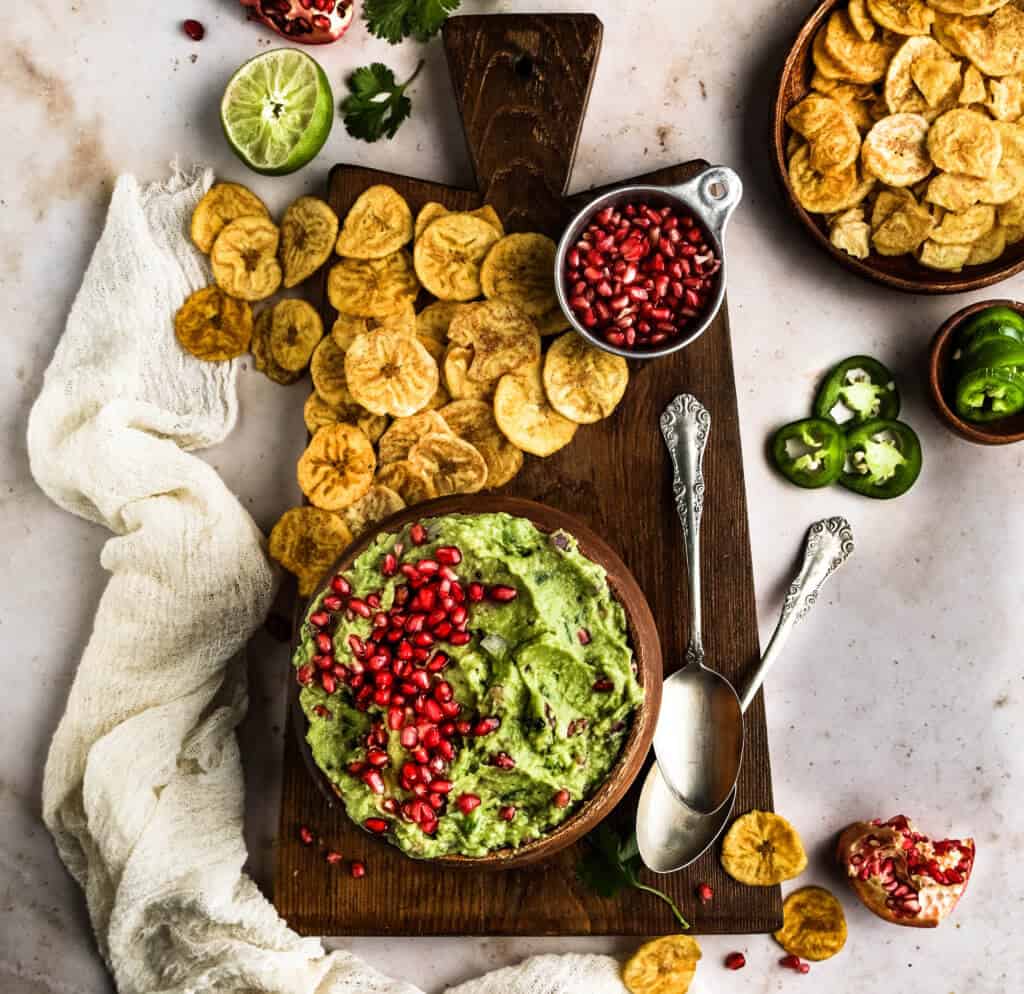 A large cutting board with  pomegranate guacamole with pomegranate aril over the top along with plantain chips scattered across the cutting board. 
