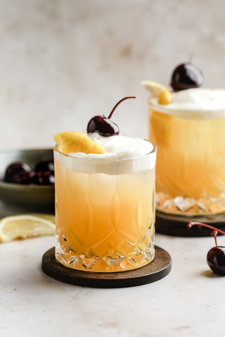 closeup of whiskey sour in a high ball glass showing the egg white foam on top with orange rind and cherry