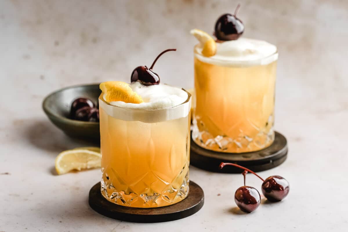 Two whiskey sour cocktails topped with a creamy egg-white foam, set on dark coasters with lemon slices and a bowl of cherries nearby on a light background.