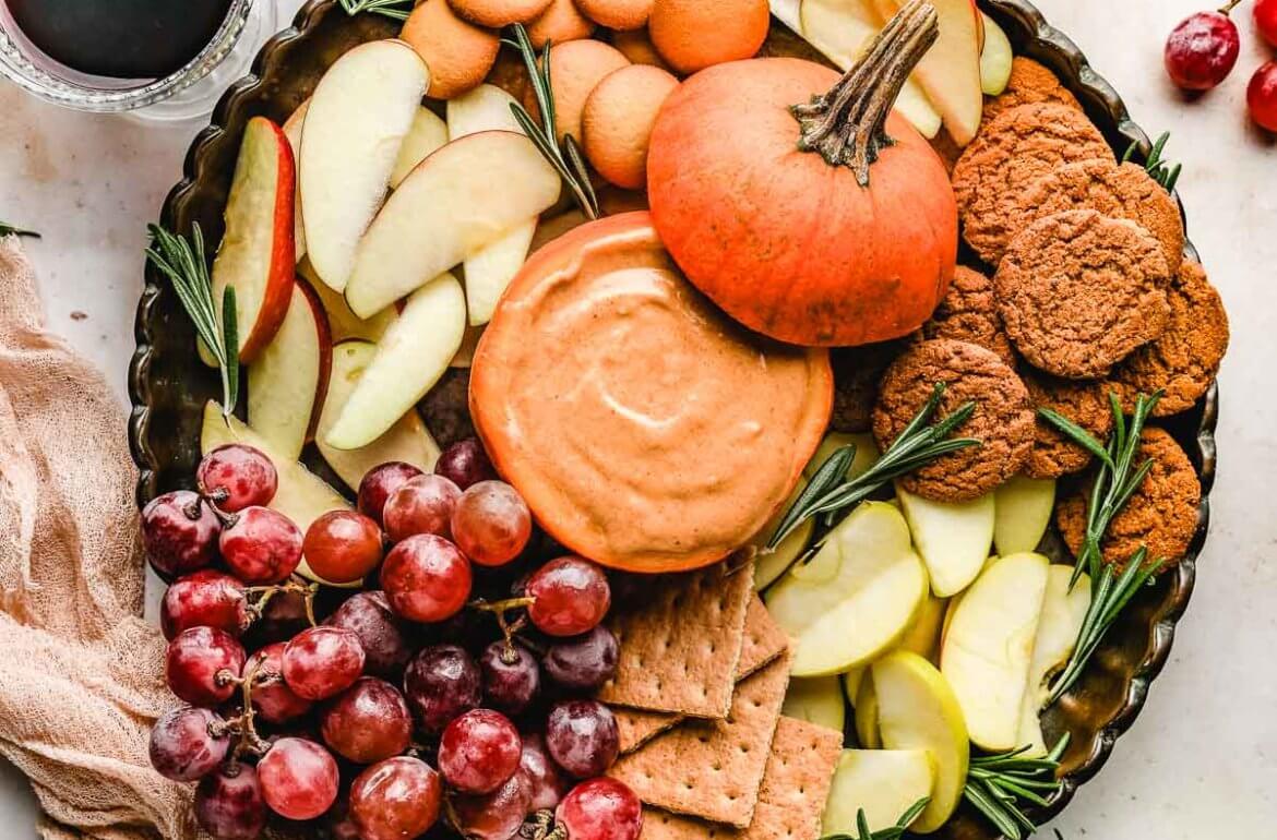 A large platter filled with pumpkin dip in a hollowed out pumpkin, grapes, apple slices, graham crackers, ginger snaps and vanilla wafers