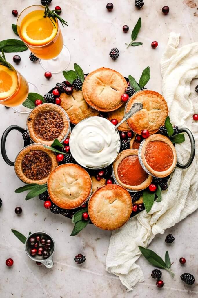 A large circle dessert charcuterie board with mini pecan, apple, blackberry and pumpkin pies served with berries and whipped cream. 