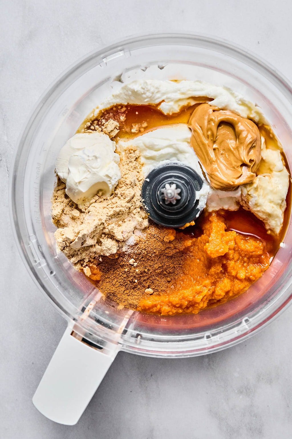 Food processor with pumpkin purée, yogurt, peanut butter, protein powder, maple syrup, vanilla, pumpkin pie spice, and salt, ready to mix.