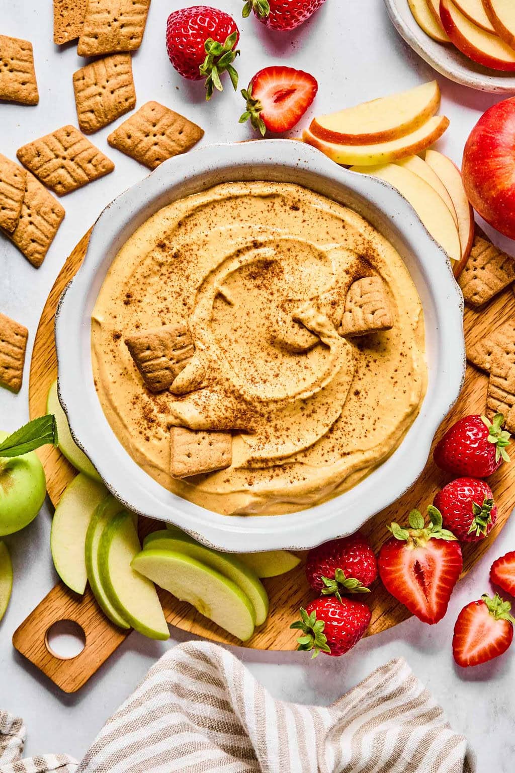 Pumpkin dip recipe in a white bowl topped with cinnamon, surrounded by apple slices, gingersnaps, strawberries, and graham crackers.