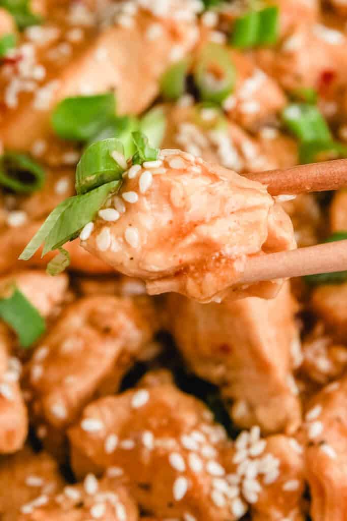 a close up photo of chopsticks holding healthy sesame chicken with sesame seeds and green onions
