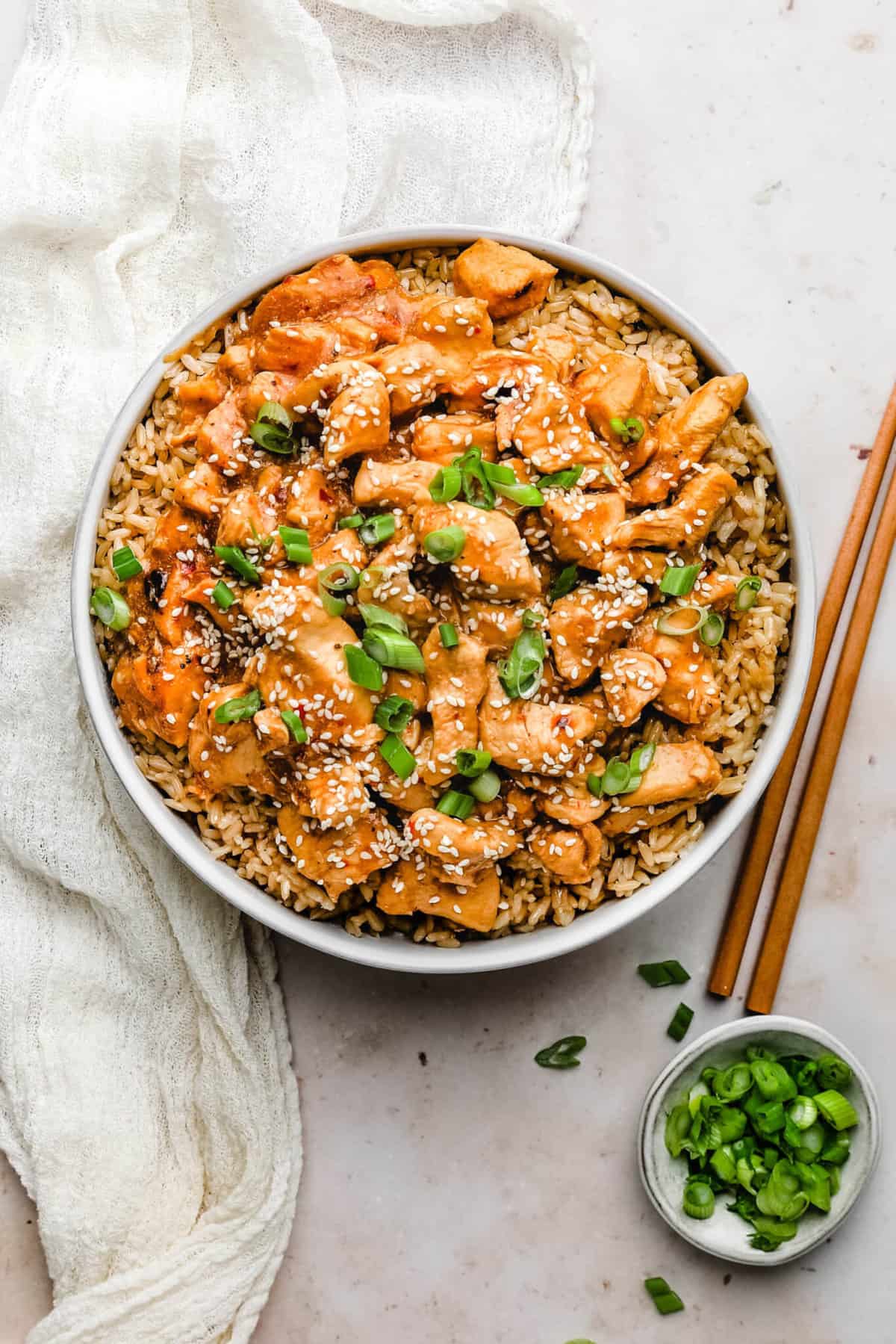 A bowl of healthy sesame chicken over brown rice garnished with green onions on a white counter top.