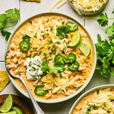 creamy dairy-free white chicken chili in a bowl filled beans and corn and topped with jalapeno, dairy-free sour cream, tortilla chips, and vegan cheese shreds