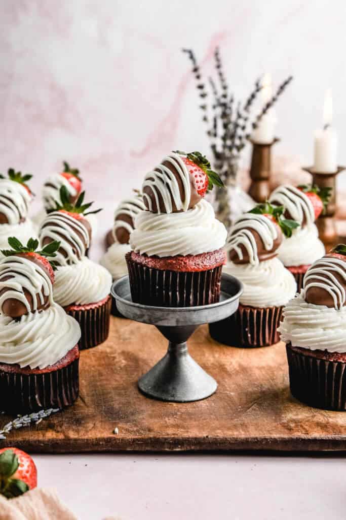 a red velvet gluten free cupcake with white buttercream icing topped with a chocolate covered strawberry on a silver stand with more red velvet cupcakes surrounding