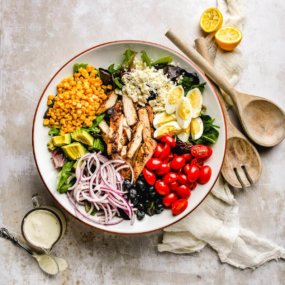 overhead shot of a healthy cobb salad that hasn't been tossed, with wooden salad spoon and fork next to it with a small cup of ranch dressing on the other side