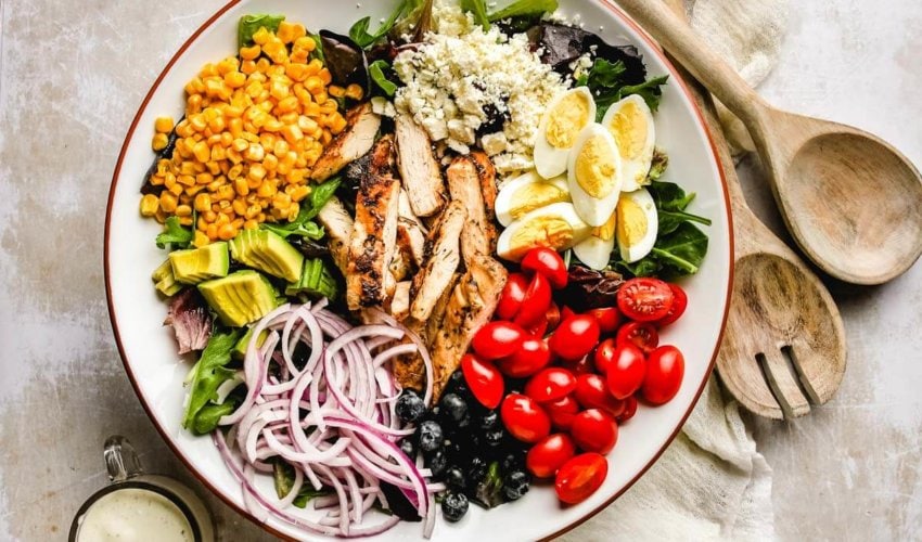overhead shot of a healthy cobb salad that hasn't been tossed, with wooden salad spoon and fork next to it with a small cup of ranch dressing on the other side