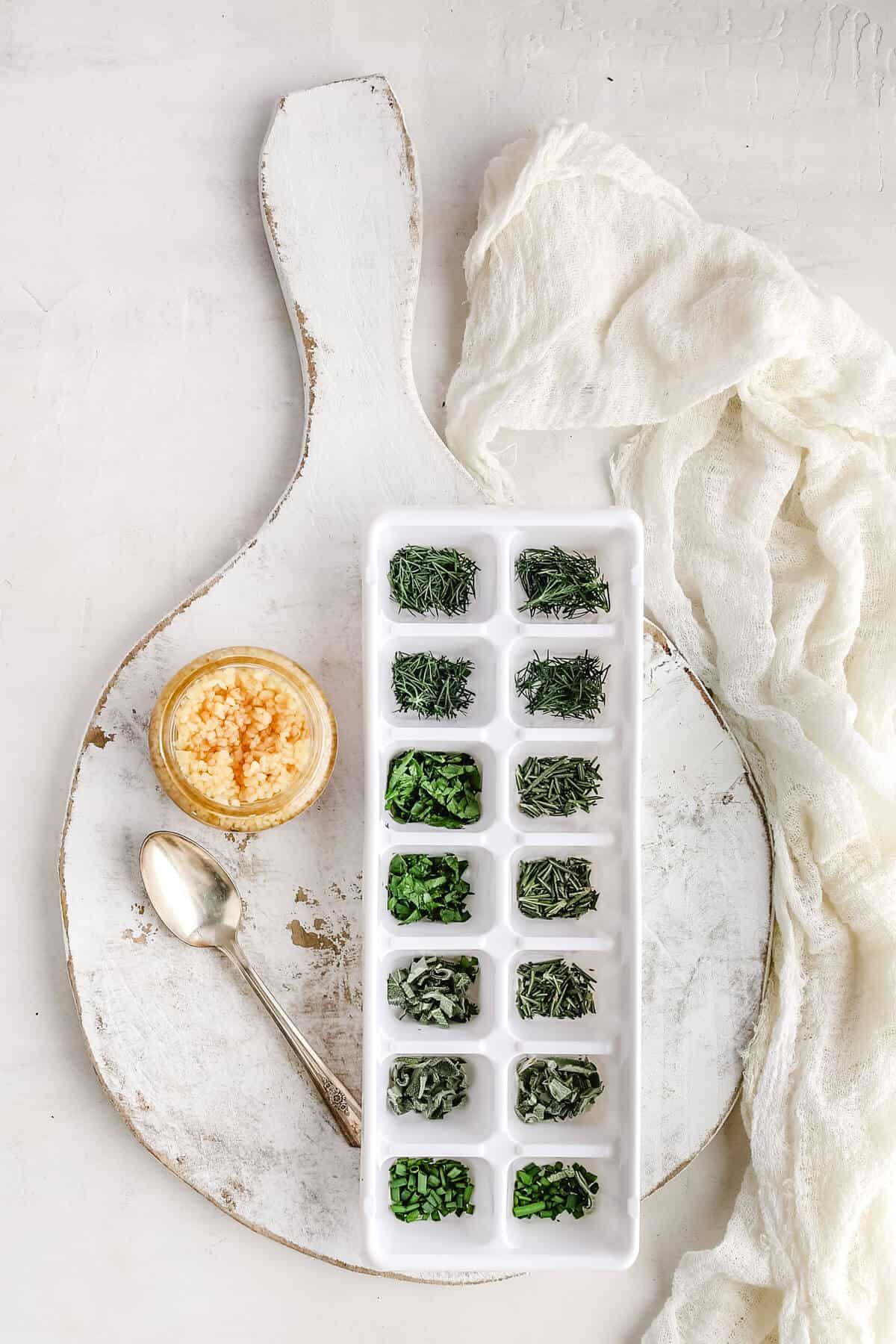 fresh herbs chopped and put into a white ice cube tray with a small bowl of garlic next to a silver spoon with a white background