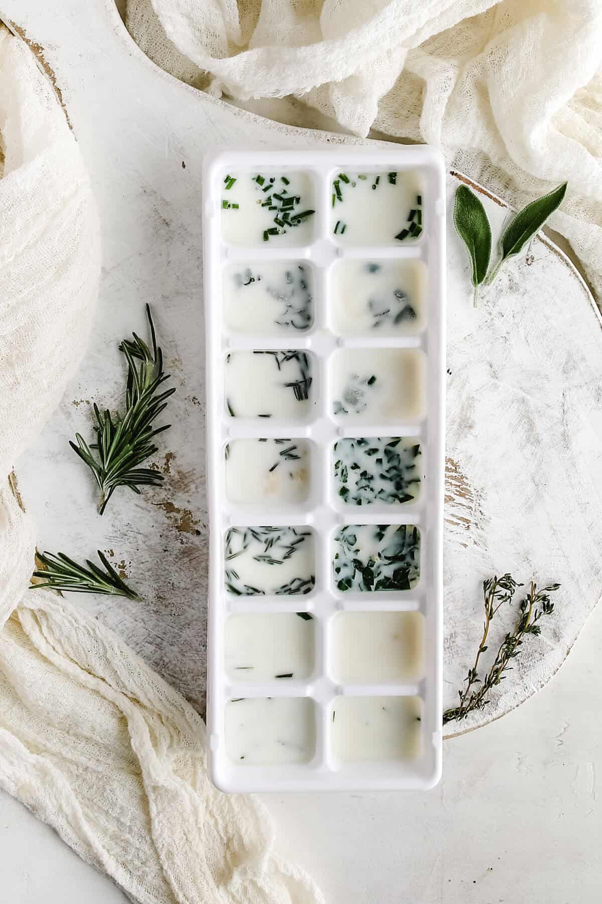 a white ice cube tray filled with fresh herbs, garlic and olive oil with sprigs of fresh herbs on a white background