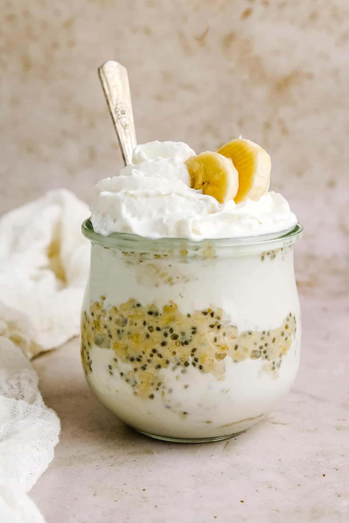 a closeup of a jar filled with layers of banana cream pie filling and chia seeds with overnight oats, topped with whipped cream and two banana slices with a silver spoon